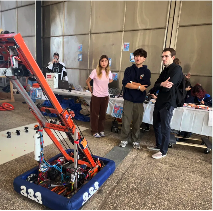 Cody Tan and Amina Akhmedova (Class of 2025 Robotics Members) are displaying MAKO —the orange robot used in the 2022–2023 CHARGED UP F.R.C Season—while engaging a curious spectator (in a black hoodie). They explain the robot's functions and the engineering behind its movement and scoring capabilities. More than just answering questions, they're fostering curiosity, inspiration, and accompanied learning that defines the core(s) of STEM outreach.